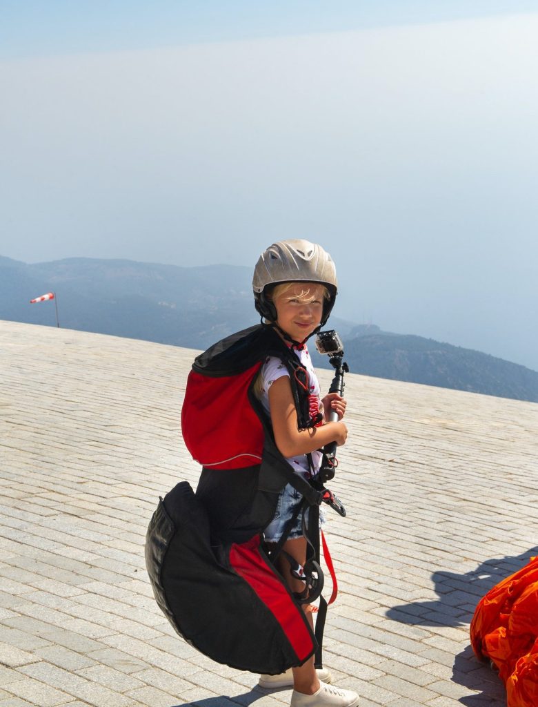 a-little-girl-in-paragliding-gear-and-with-a-camera-stands-on-the-edge-of-mount-babadag-turkey-e1655171742105.jpg